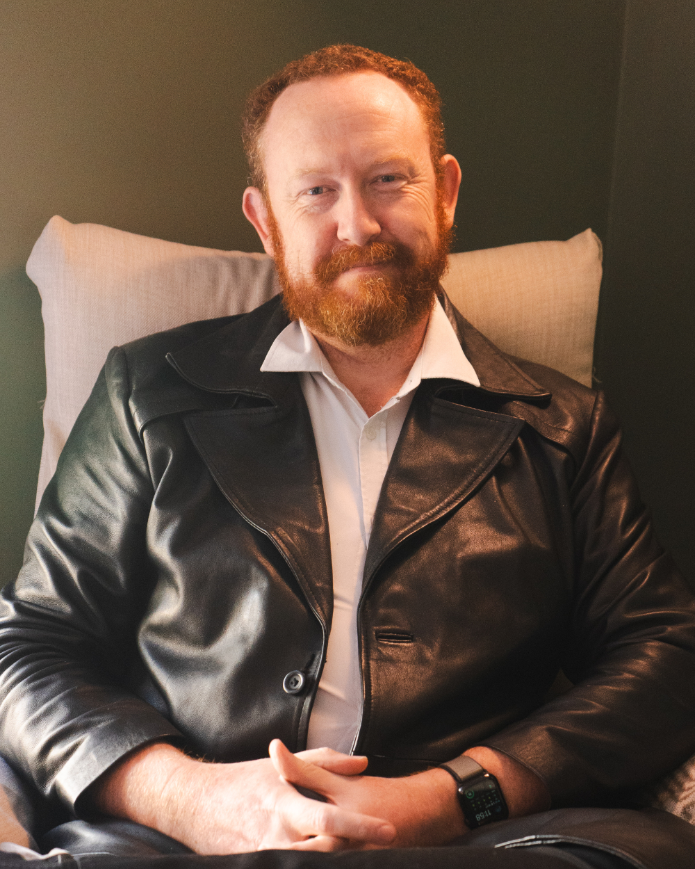 A man with red hair sitting in a chair. He is smiling, with his hands clasped in his lap.