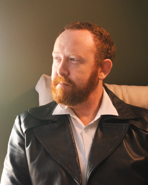 A portrait of a red headed man in afternoon light. Wearing a white button down shirt and leather jacket, He looks pensive, smiling slightly.
